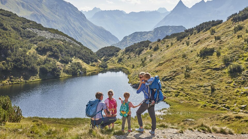 A family is hiking in the mountains next to a clear lake. The landscape is green and hilly, surrounded by impressive peaks.