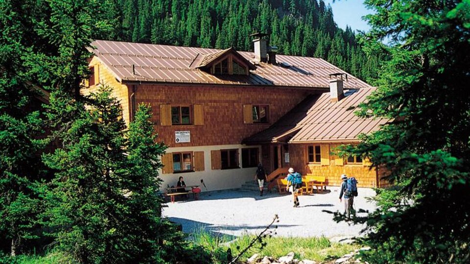A mountain cabin surrounded by tall fir trees. In the foreground, hikers can be seen enjoying nature.