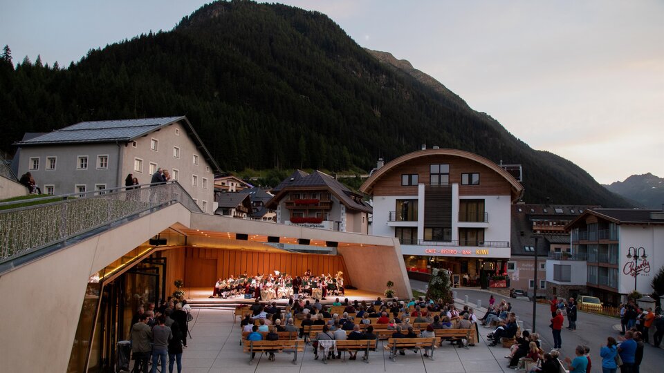 An outdoor event with an orchestra is playing against a backdrop of mountains. Many people are sitting on benches and enjoying the music.