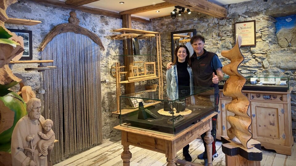 A woodworking shop with displayed sculptures and a workshop. Two people are standing in the middle of the room and smiling.