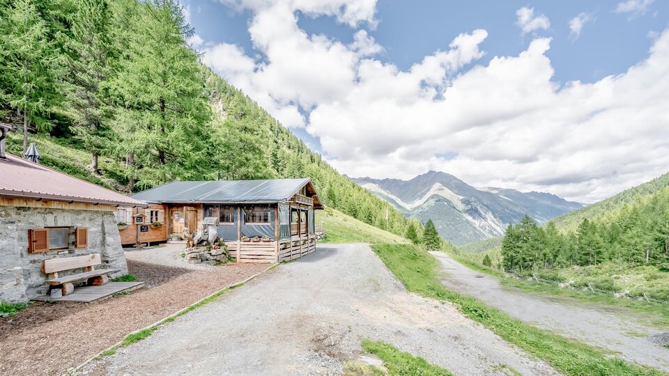 A picturesque Alpine landscape with two cottages and green trees. The mountains in the background are visible under a clear sky.