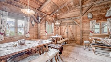 A cozy wooden room with a large table and benches. The walls are made of wood, and through the windows, you can see the nature outside.