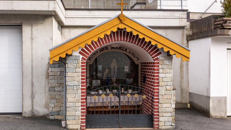 A small chapel with a yellow roof and a cross. It is built of stone and bricks and has a closed lattice door. | © Bernhard Gruber – Photography
