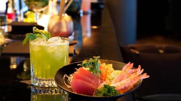 A delicious cocktail with fresh herbs stands next to a plate of sushi and seafood. The colorful presentation on an elegant table invites enjoyment.