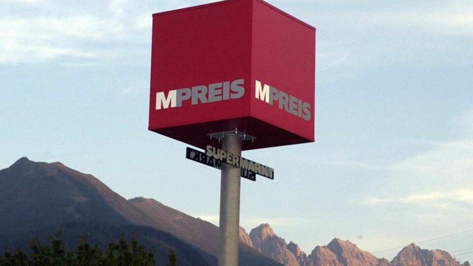 Ein rotes Geschäftsschild mit der Aufschrift "MPREIS" vor einem Bergpanorama. Der Himmel ist klar und die Landschaft ist grün.