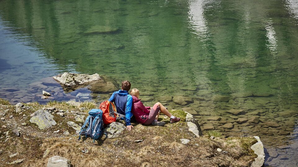 A peaceful landscape by the shore of a clear lake. Two people are sitting relaxed on the stones and enjoying the view.