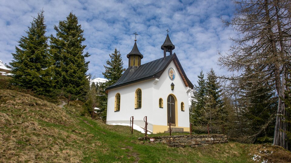 A small, charming chapel stands on a hill, surrounded by green meadows and tall fir trees. The sky is clear and shows some clouds. | © Bernhard Gruber – Photography