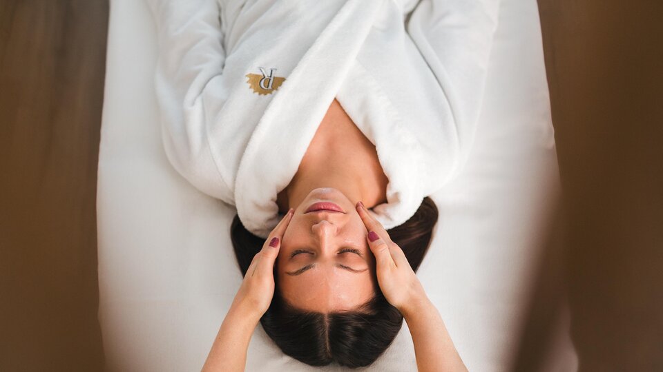 A woman is lying relaxed on a lounger receiving a facial massage. She is wearing a white bathrobe and looks happy.