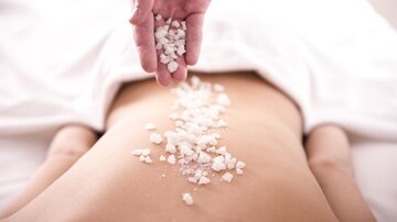 A hand sprinkles salt on the back of a lying person. It symbolizes relaxation and wellness.
