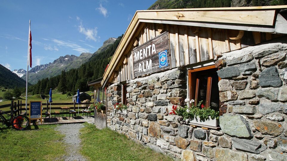 A cozy stone mountain hut in a picturesque mountain landscape. The surroundings are green, with mountains in the background and blue sky.