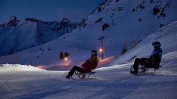Two people are sitting in armchairs on a snowy area under an evening sky. In the background, mountains and a ski lift cable car can be seen.