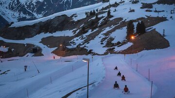 A wintry mountain landscape with snow-covered mountains and a slope in the foreground. Some people are sledding down the hill as twilight sets in.