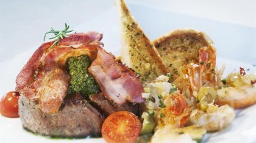 A tender steak, wrapped in crispy bacon and served with shrimp, tomatoes, and toasted bread. The dish is elegantly presented and looks appetizing.