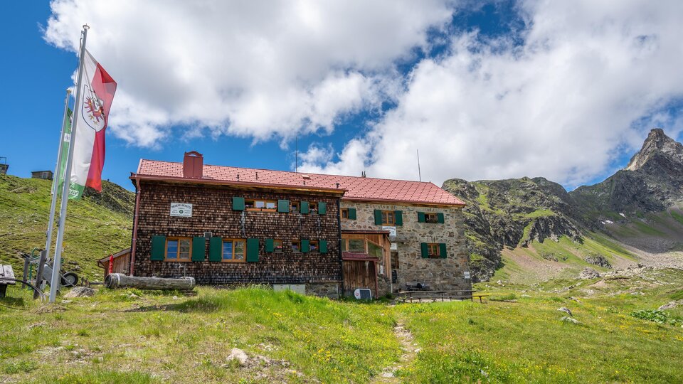 A stone mountain hut in the midst of a green meadow. In the background, mountains and a blue sky with some clouds can be seen.