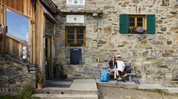 A cozy stone mountain cabin with green shutters. A couple sits relaxed in front of the cabin with a bag next to them.