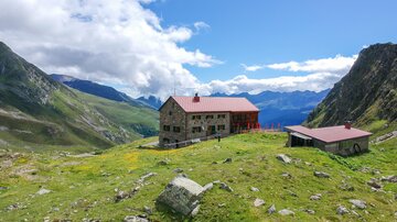 A mountain cabin surrounded by green meadows and majestic mountains. The sky is clear with a few clouds.