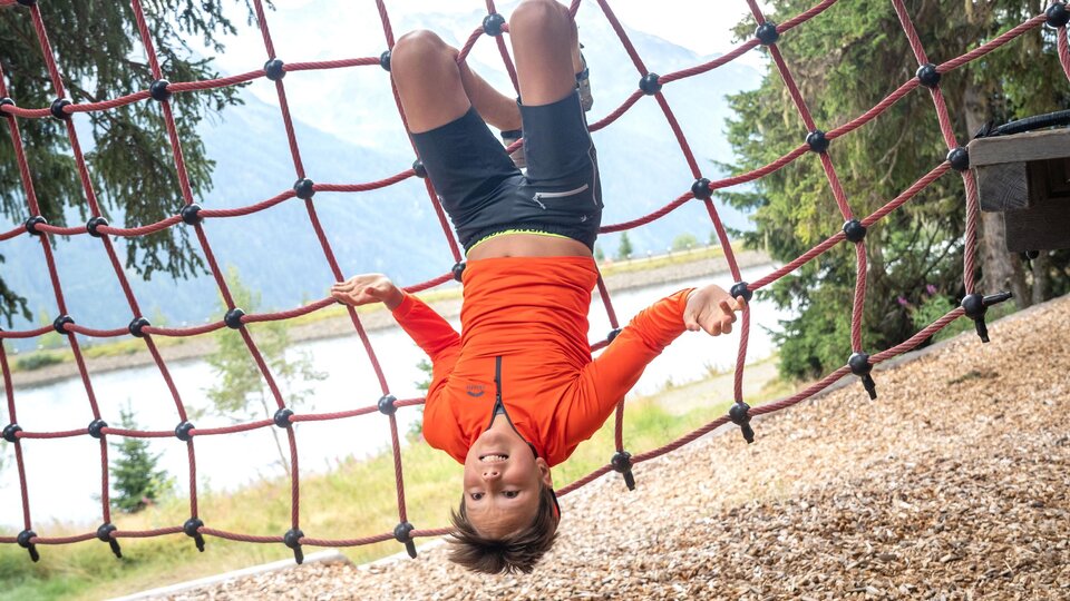 A boy is hanging upside down in a red climbing net and is happily looking at the camera. In the background, there are trees, a lake, and the blurred mountain landscape. The image captures a playful moment full of energy, courage, and lightness in the alpine outdoor experience.