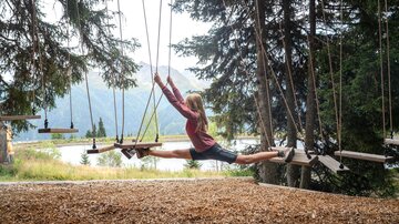 A girl balances with outstretched legs between two floating wooden elements of a low rope garden. Behind her, the view opens up to a mountain lake and the surrounding alpine landscape. The scene appears sporty, free, and adventurous, showcasing nature experience and dexterity amidst the Paznaun mountain world.