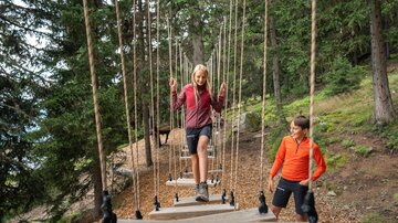 Two children are exploring a ropes course in the forest amidst dense spruces. A girl is balancing intently on suspended wooden planks, while a boy in orange sportswear stands next to her, smiling at her. The natural setting with forest floor, trees, and alpine backdrop conveys adventure, movement, and family time in nature.