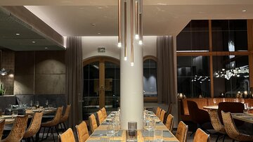 An elegant restaurant with a long wooden table and stylish chairs. The lighting creates a cozy atmosphere.