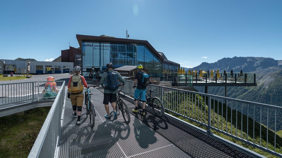 Three cyclists are standing on an observation platform overlooking the mountains. In the background, a modern building can be seen.
