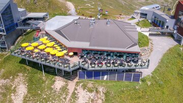 A modern restaurant on a mountain with a terrace and many sun umbrellas. The surroundings are green and mountainous, ideal for a relaxing stay.