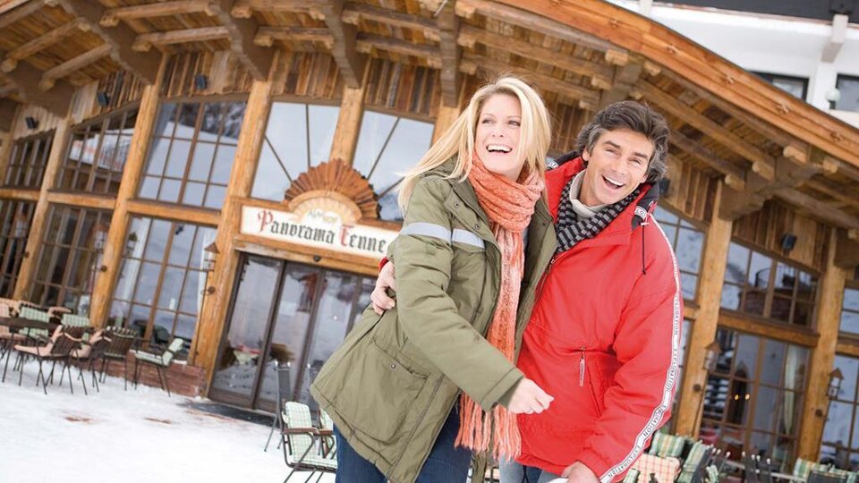 A happy couple stands smiling in front of a wooden building in the snow. In the background, there is a terrace with chairs and tables visible.