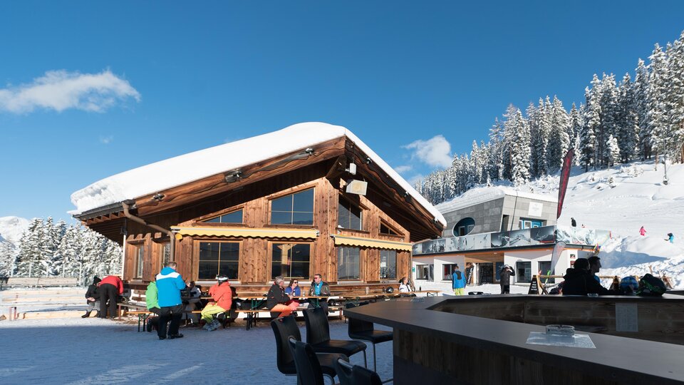 A cozy mountain cabin in the snow with guests outside. Snow-covered mountains and a blue sky are visible in the background.
