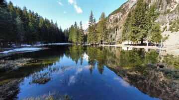 A calm lake surrounded by tall trees and mountains. The smooth water surface reflects the landscape.