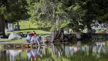 A group of people sits by the shore of a quiet pond, surrounded by green trees. The water reflects the landscape and the group.