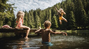 Three children are playing by the shore of a lake. One child joyfully jumps into the water while the others watch.