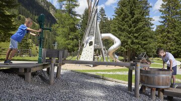A playground in nature with various play equipment. Two children are actively playing with a water pump and a barrel.
