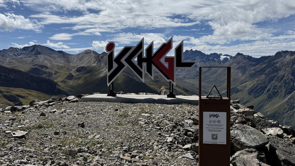 A mountain peak with a large sign that says "Ischgl". In the background, majestic mountains and a clear sky are visible.