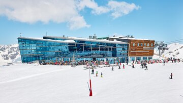 Eine moderne Bergstation umgeben von Schnee und Bergen. Viele Menschen genießen die Aktivitäten im Freien.