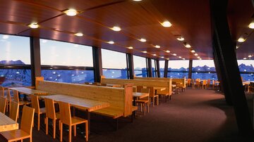 Ein modernes Restaurant mit großen Fenstern und einem Blick auf die Berge. Die Einrichtung ist schlicht mit Holz-Tischen und -Stühlen.