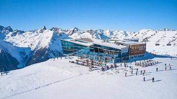 Ein modernes Bergrestaurant in den Alpen umgeben von schneebedeckten Bergen. Skifahrer genießen die sonnige Winterlandschaft.