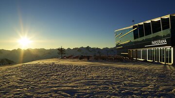 Ein modernes Gebäude steht in einer schneebedeckten Landschaft. Im Hintergrund sieht man die Berge und die Sonne, die den Himmel erhellt.