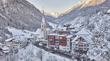A picturesque winter village with snow-covered mountains and a church in the foreground. The streets are quiet, surrounded by white fir trees and charming houses.
