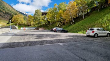 Ein Parkplatz in einer malerischen Landschaft umgeben von Bäumen und Bergen. Der Himmel ist blau mit ein paar Wolken.