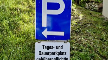 Ein Parkplatzschild mit dem Hinweis, dass das Parken gebührenpflichtig ist. Es zeigt die Richtung nach links.