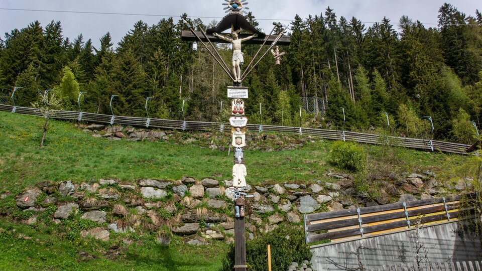 An artistic wooden Stations of the Cross featuring various figures and symbols. In the background, a green meadow and wooded hills stretch out. | © Bernhard Gruber – Photography