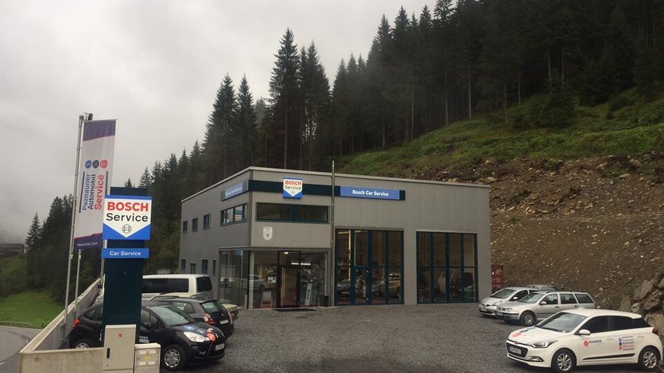 A modern service building by Bosch, surrounded by forests and mountains. Several cars are parked in the parking lot.
