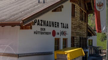 A traditional mountain hut sign with the inscription "Paznauner Taja - the hut meeting place in Ischgl". The hut is made of wood and surrounded by green mountains.