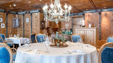 An elegant restaurant with a round table and blue chairs. Above the table hangs a sparkling chandelier in a room paneled with wood.