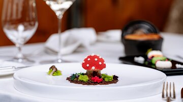 An artfully arranged dish with a red chocolate mushroom placed on a bed of colorful vegetables and herbs. In the background, glasses of wine and other dishes are visible.
