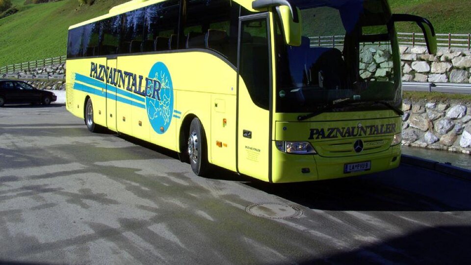 A yellow tour bus is parked in a parking lot in a mountainous landscape. In the background, green hills and some houses are visible.