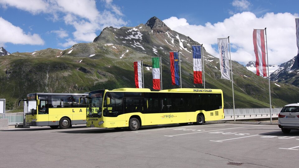 Two yellow buses are parked in a parking bay in front of an impressive mountain landscape. In the background, flags of various countries flutter in the clear sky.