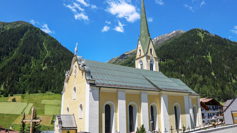 A picturesque church with a tall tower and a yellow facade, surrounded by green mountains. The clear sky makes the landscape appear radiant.