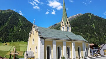 A picturesque church with a tall tower and a yellow facade, surrounded by green mountains. The clear sky makes the landscape appear radiant.