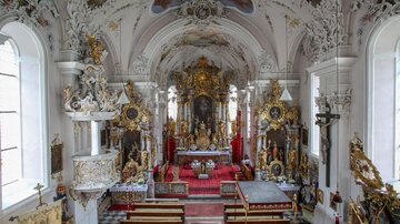 A magnificent church with an ornate ceiling and many decorations. Benches are arranged in rows, and the altar is at the center of the room.